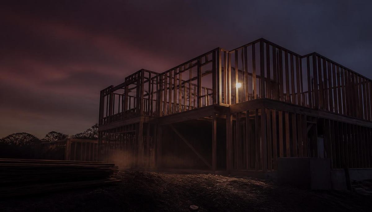 Timber wall framing of a two-storey Australian home at dusk, lit by a single work light against a deep indigo sky